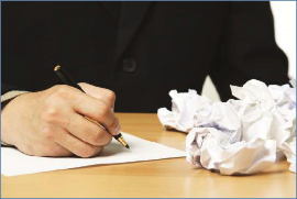 
The photo shows a  person writing with a pen, surrounded by crumpled paper from previous attempts.