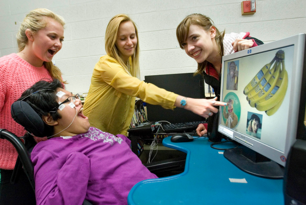 Image of student using EagleEyes to play a game in the computer as she's laughing. The teacher is pointing to an icon on the computer screen while two other females are smiling too. 