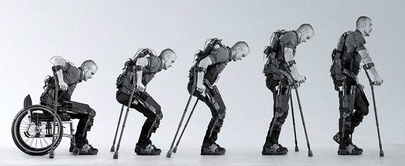Picture shows a man sitting in a wheelchair as he put on the exoskeleton suit it shows five level of the man standing up as he slowly gain his stands while using a walker he is able to balance himself and starts moving forward as he walks.