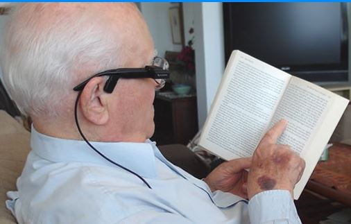 Shows an older gentalmen sitting down, as he is holding up his book and pointing to the page to show the OrCam where to start reading. the OrCam is already placed on the side of his glasses.